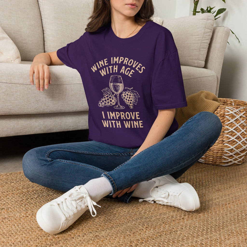 Person wearing a purple t-shirt with text "Wine Improves With Age. I Improve With Wine" in an off-white bold font. Image of wine glass and grape bunches separate text sitting on the floor.