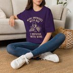 Person wearing a purple t-shirt with text "Wine Improves With Age. I Improve With Wine" in an off-white bold font. Image of wine glass and grape bunches separate text sitting on the floor.