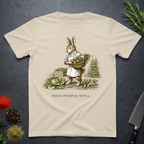 Sand colored t-shirt with a printed design of a rabbit holding a basket of vegetables and the text 'Feed People Well' on a dark surface.