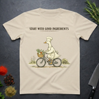 Sand colored t-shirt with a graphic of a goose chef riding a bike with vegetables in basket. Text "Start with Good Ingredients" above graphic. 