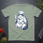 Sage green t-shirt with a navy and white Cheshire chef cat graphic holding a knife on a dark surface.