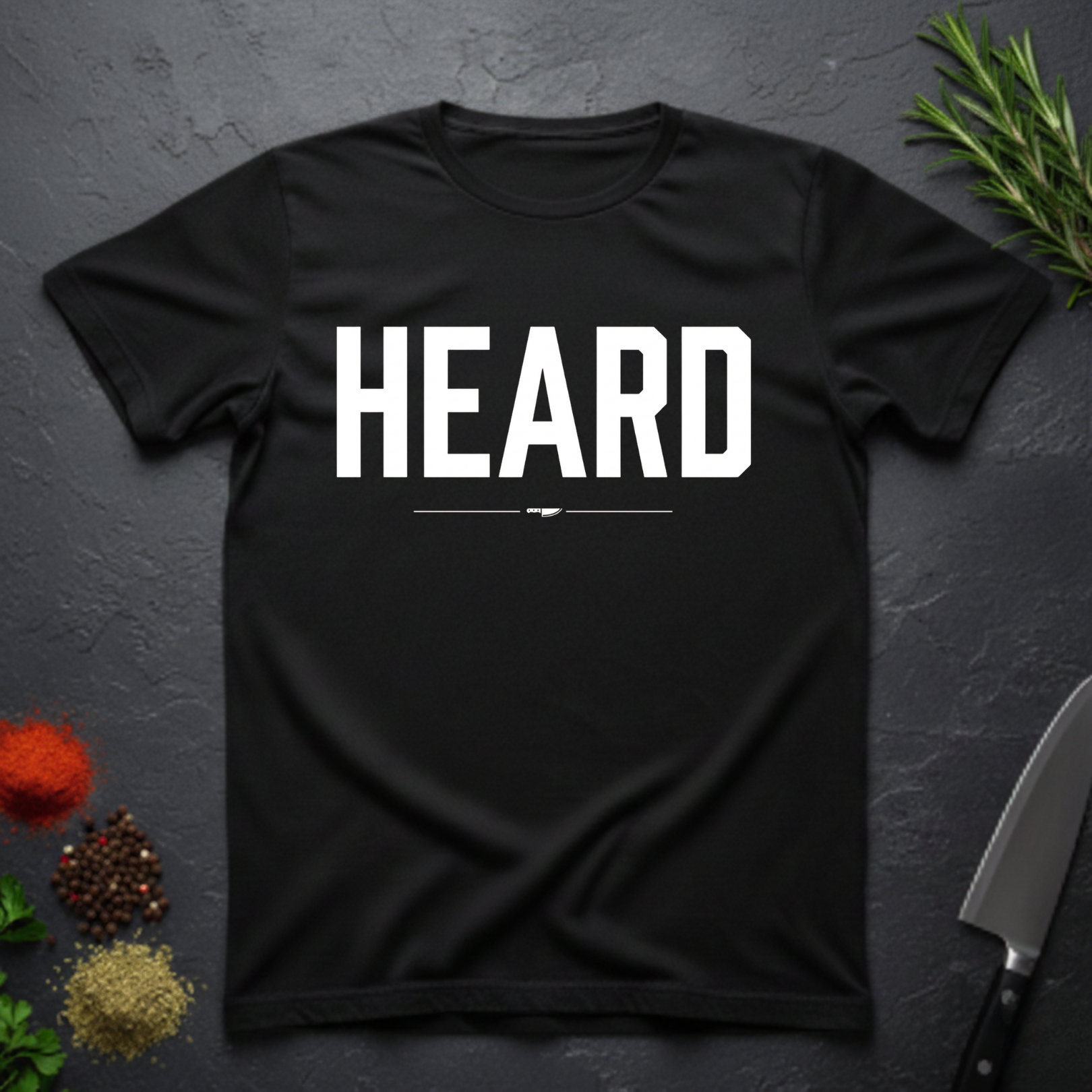 Black t-shirt with 'HEARD' in bold white text, placed on a dark surface with decorative elements.