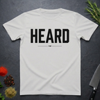 Ice Grey  t-shirt with 'HEARD' in bold black text, placed on a dark surface with decorative elements.