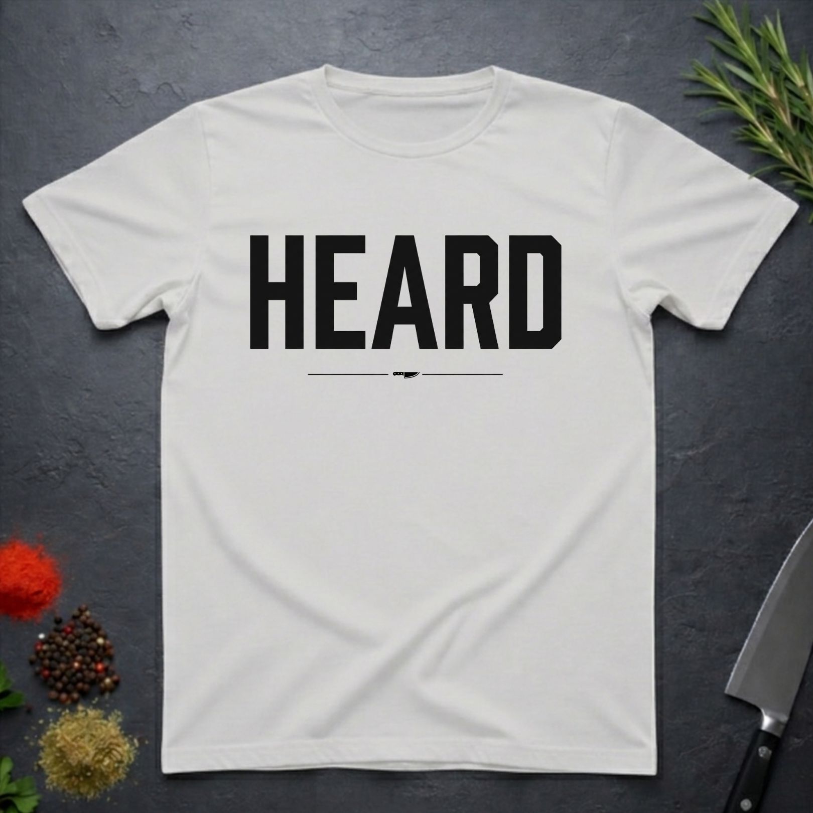 Ice Grey  t-shirt with 'HEARD' in bold black text, placed on a dark surface with decorative elements.