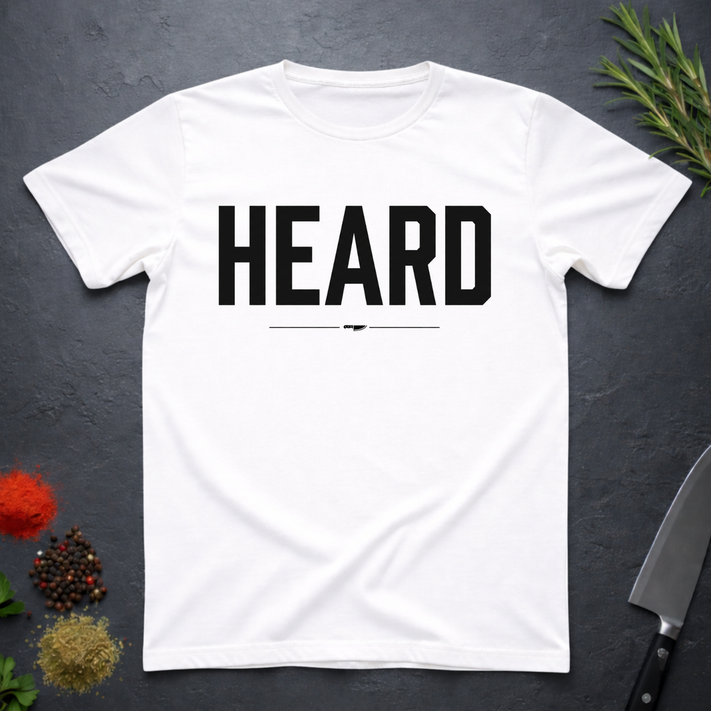 White t-shirt with 'HEARD' in bold black text, placed on a dark surface with decorative elements.