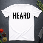 White t-shirt with 'HEARD' in bold black text, placed on a dark surface with decorative elements.