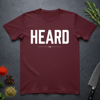 Maroon t-shirt with 'HEARD' in bold white text, placed on a dark surface with decorative elements.