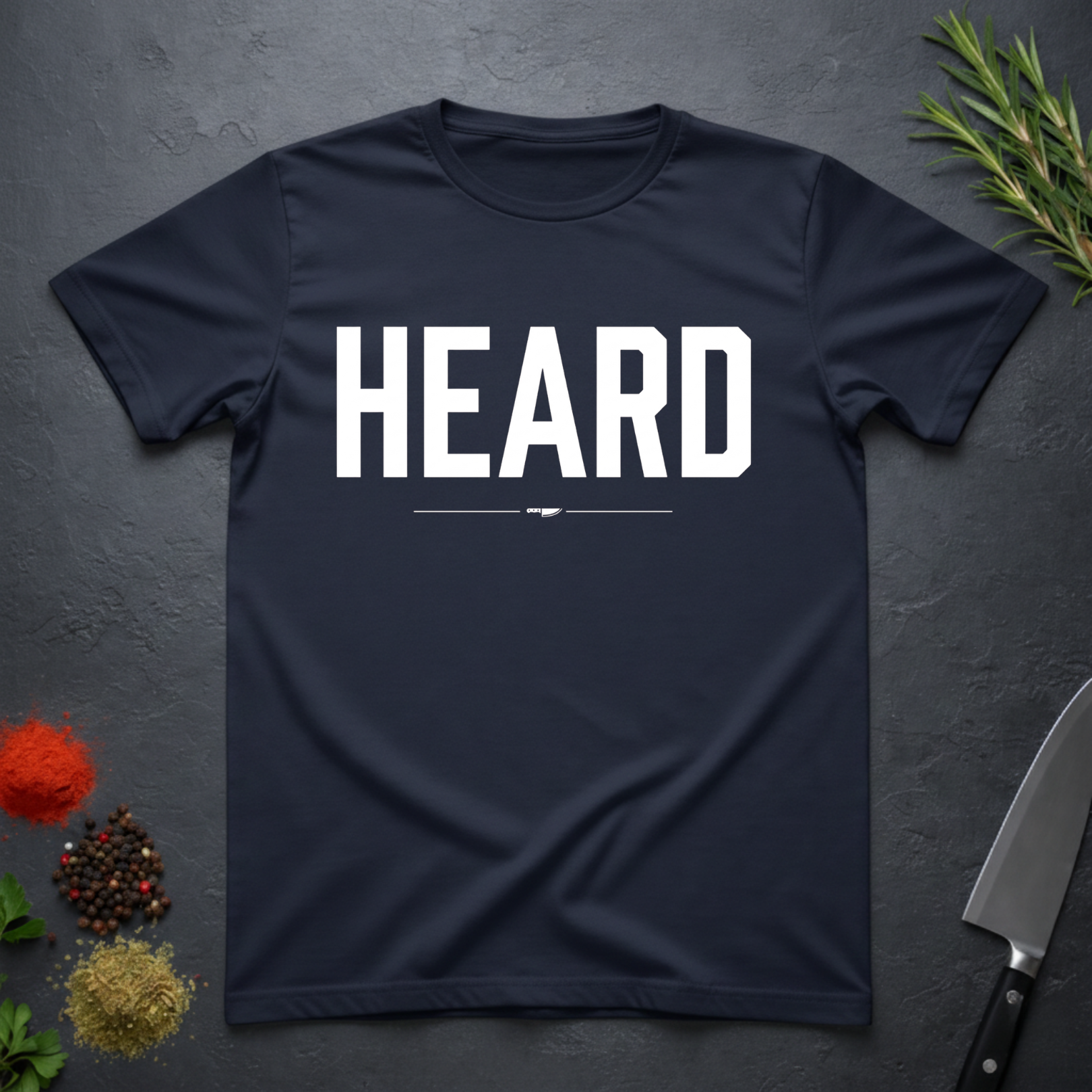 Navy Blue t-shirt with 'HEARD' in bold white text, placed on a dark surface with decorative elements.