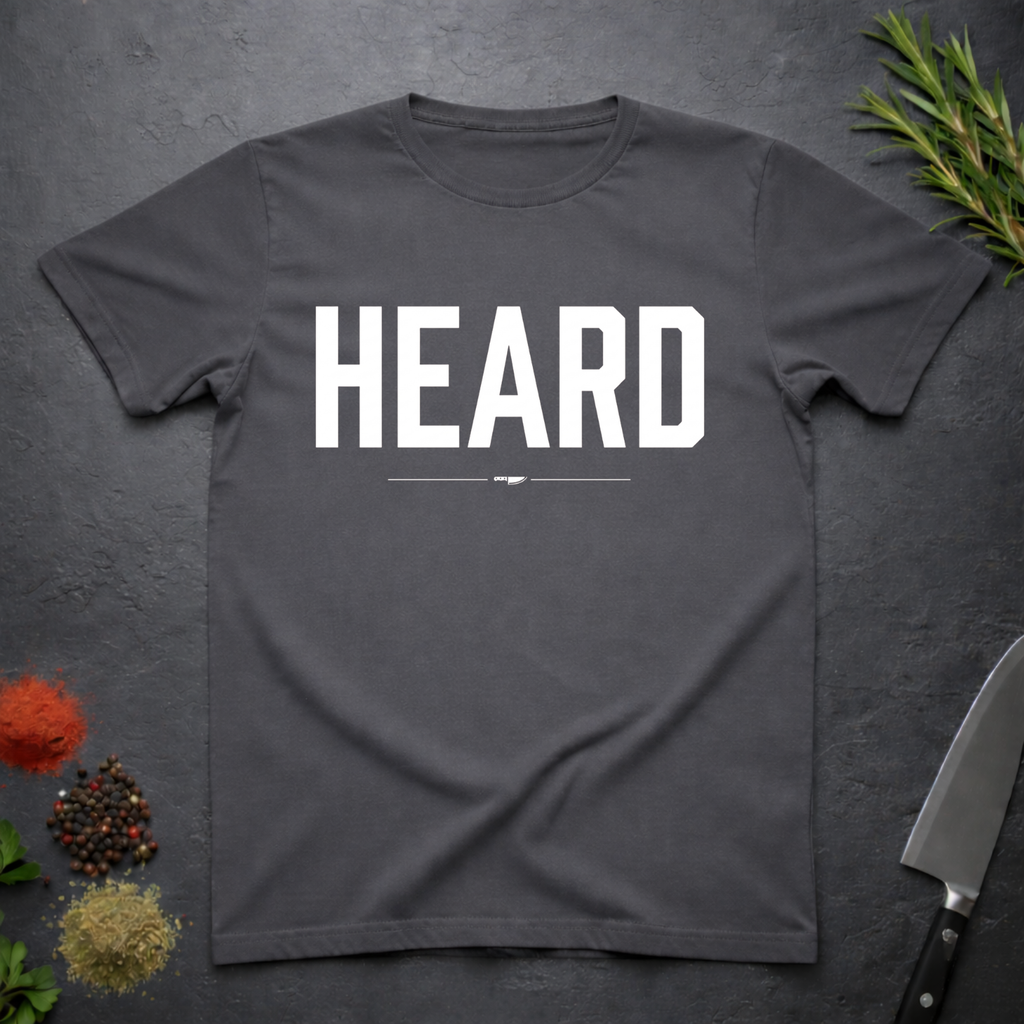 Charcoal Grey t-shirt with 'HEARD' in bold white text, placed on a dark surface with decorative elements.