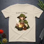 Sand colored t-shirt with a anime type elf character cooking over a fire, text 'Cook with Magic', on a dark surface with kitchen items.
