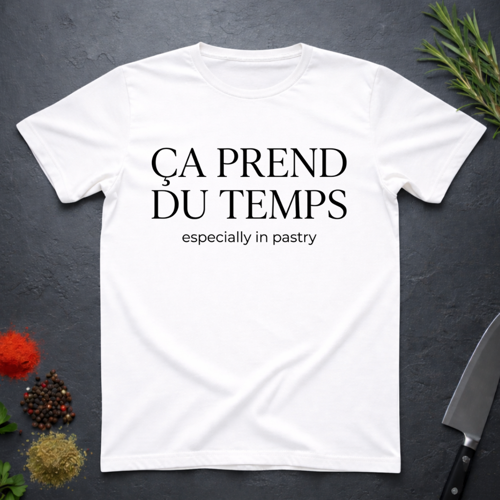 White t-shirt with text 'Ca prend du temps especially in pastry' on a dark surface with kitchen items.