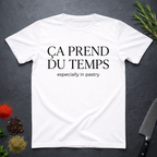 White t-shirt with text 'Ca prend du temps especially in pastry' on a dark surface with kitchen items.