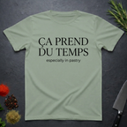 Sage  green t-shirt with text 'Ca prend du temps especially in pastry' on a dark surface with kitchen items.