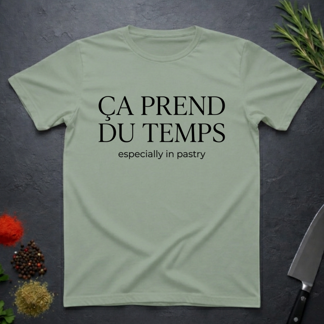 Sage  green t-shirt with text 'Ca prend du temps especially in pastry' on a dark surface with kitchen items.