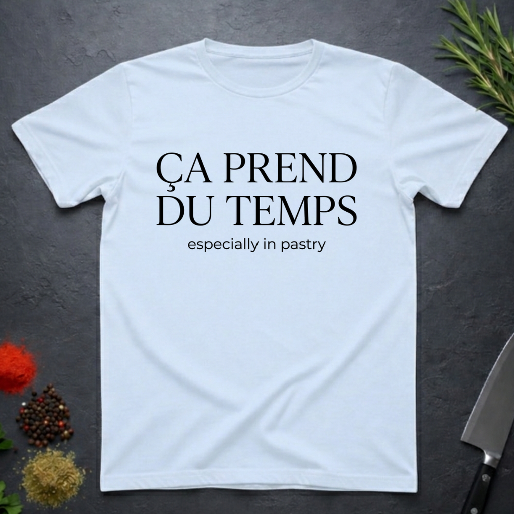 Light Blue t-shirt with text 'Ca prend du temps especially in pastry' on a dark surface with kitchen items.