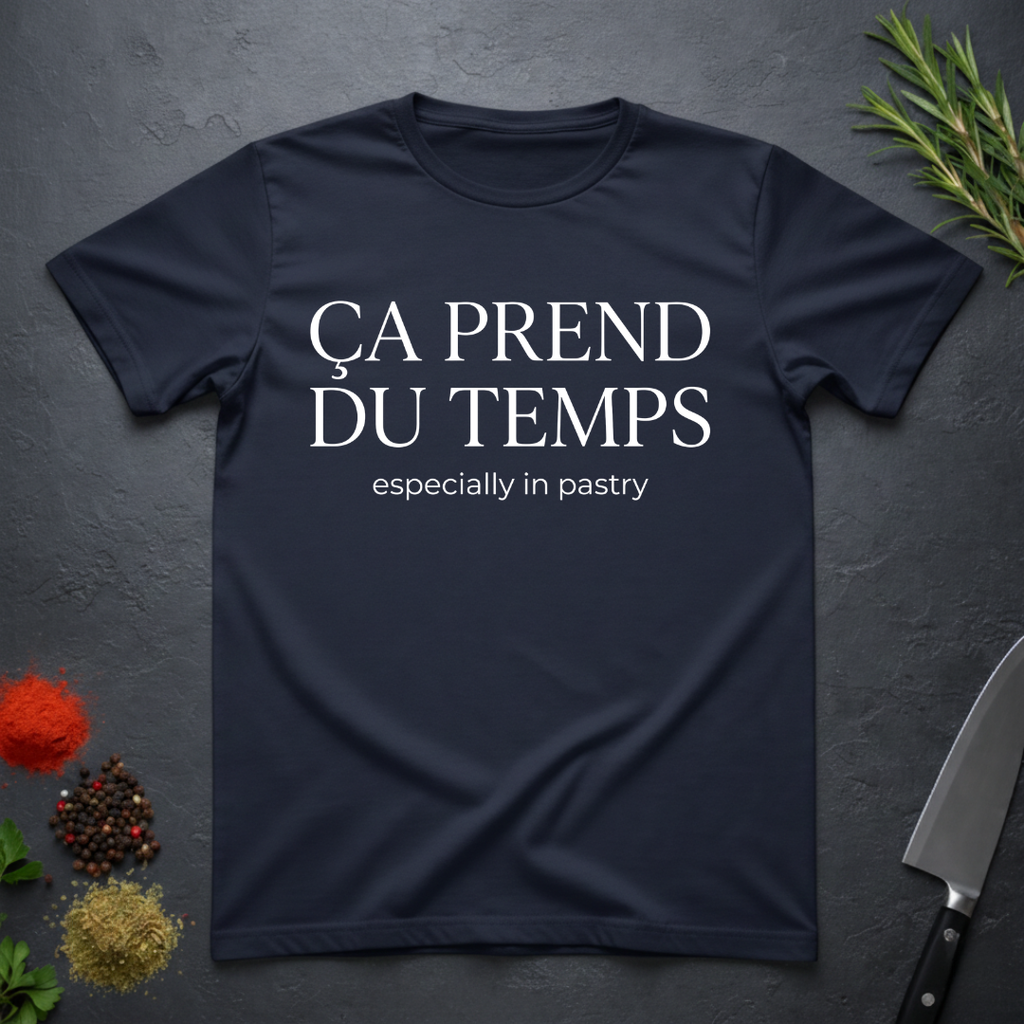Navy blue t-shirt with text 'Ca prend du temps especially in pastry' on a dark surface with kitchen items.