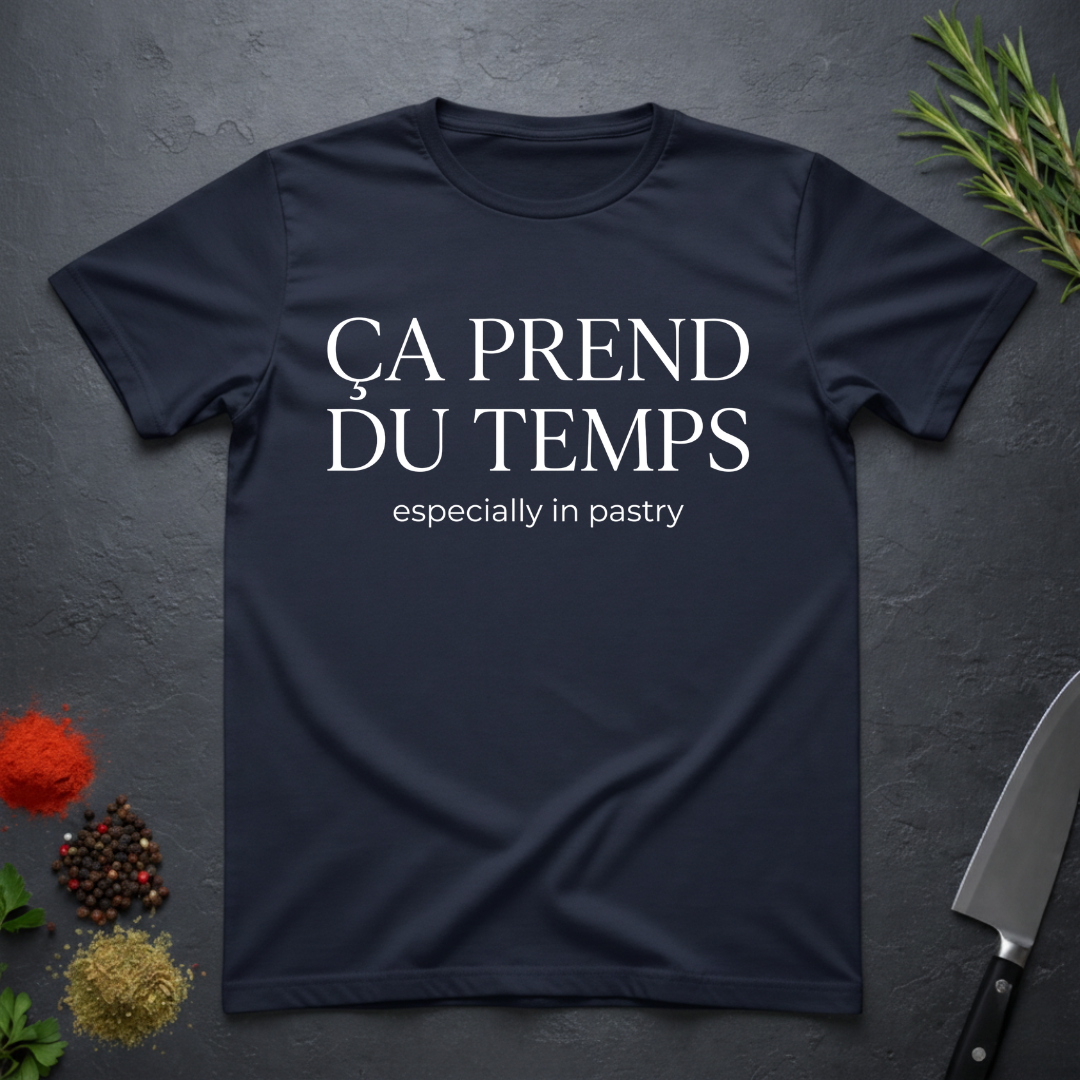 Navy blue t-shirt with text 'Ca prend du temps especially in pastry' on a dark surface with kitchen items.