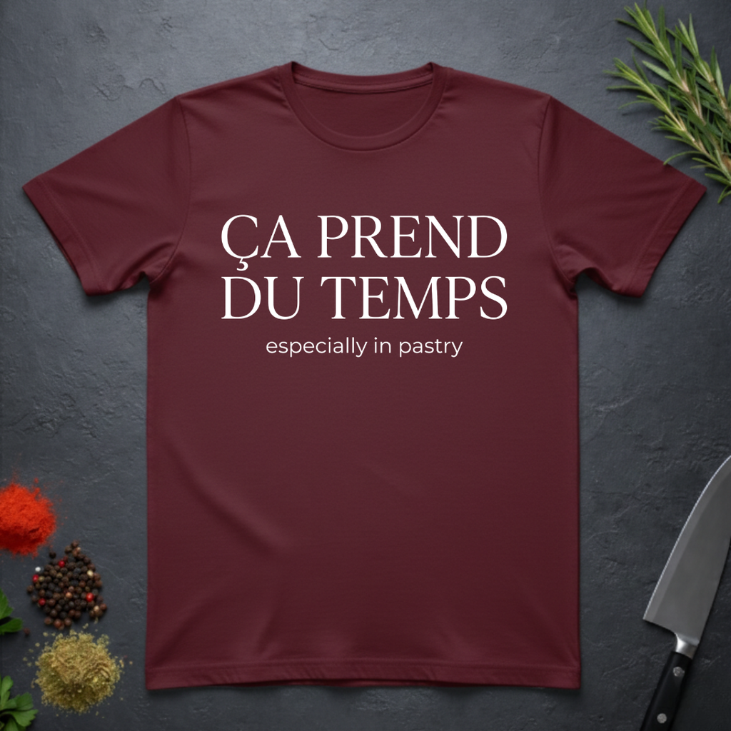 Maroon t-shirt with text 'Ca prend du temps especially in pastry' on a dark surface with kitchen items.