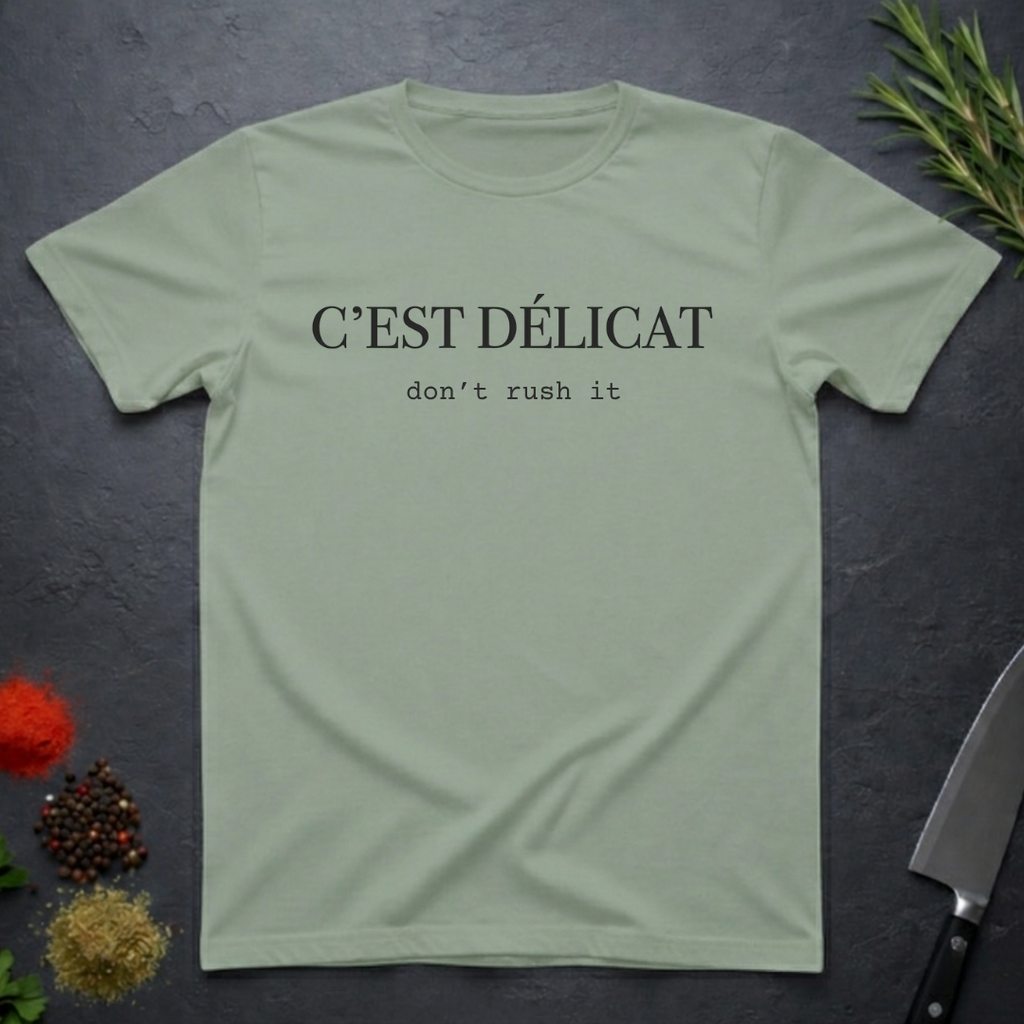 Sage colored t-shirt with text 'C'EST Délicat don't rush it' on a dark surface with decorative elements.