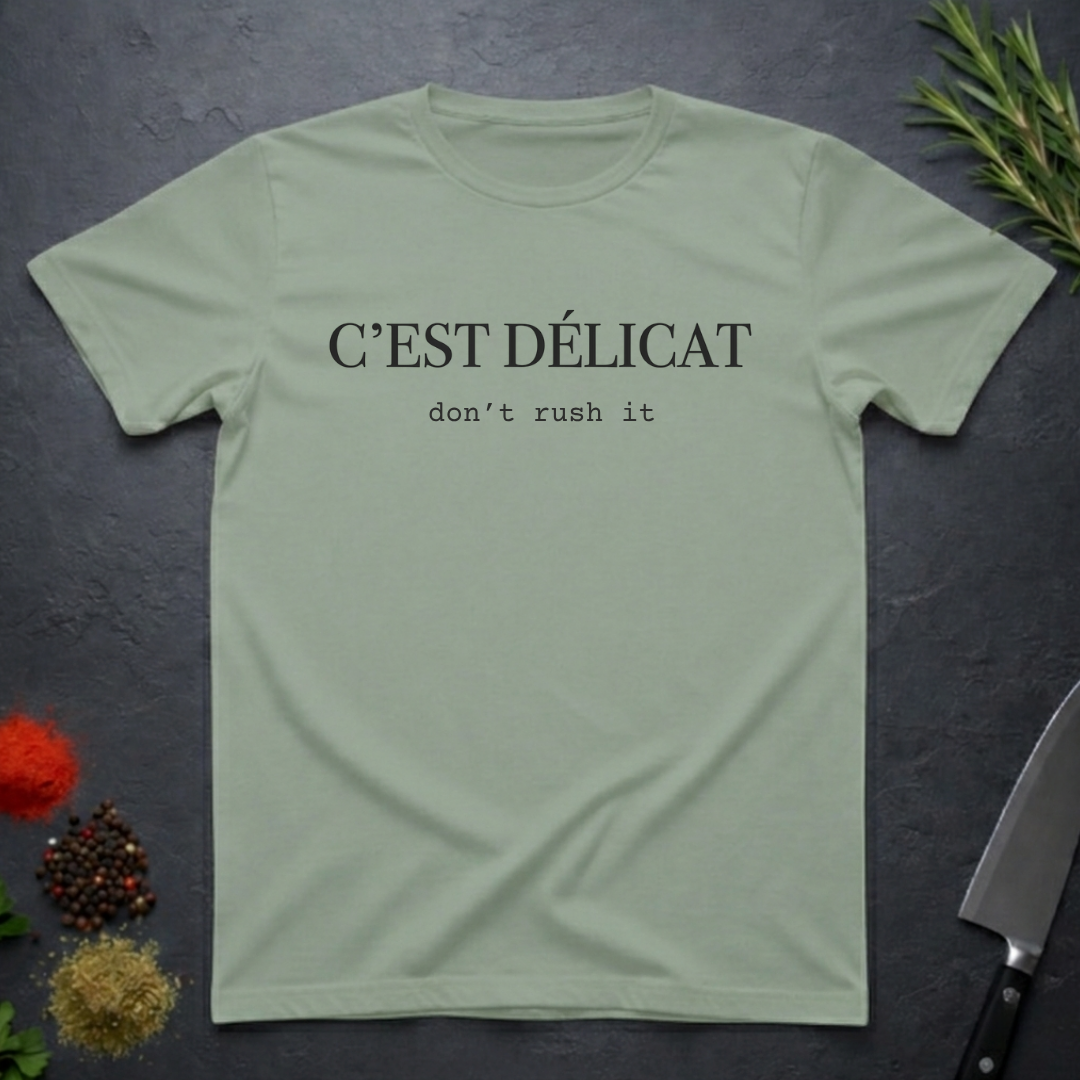 Sage colored t-shirt with text 'C'EST Délicat don't rush it' on a dark surface with decorative elements.