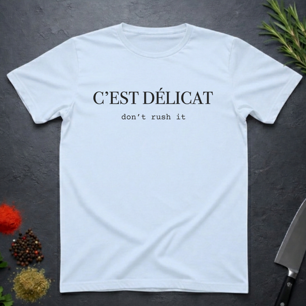 Light Blue t-shirt with text 'C'EST Délicat don't rush it' on a dark surface with decorative elements.