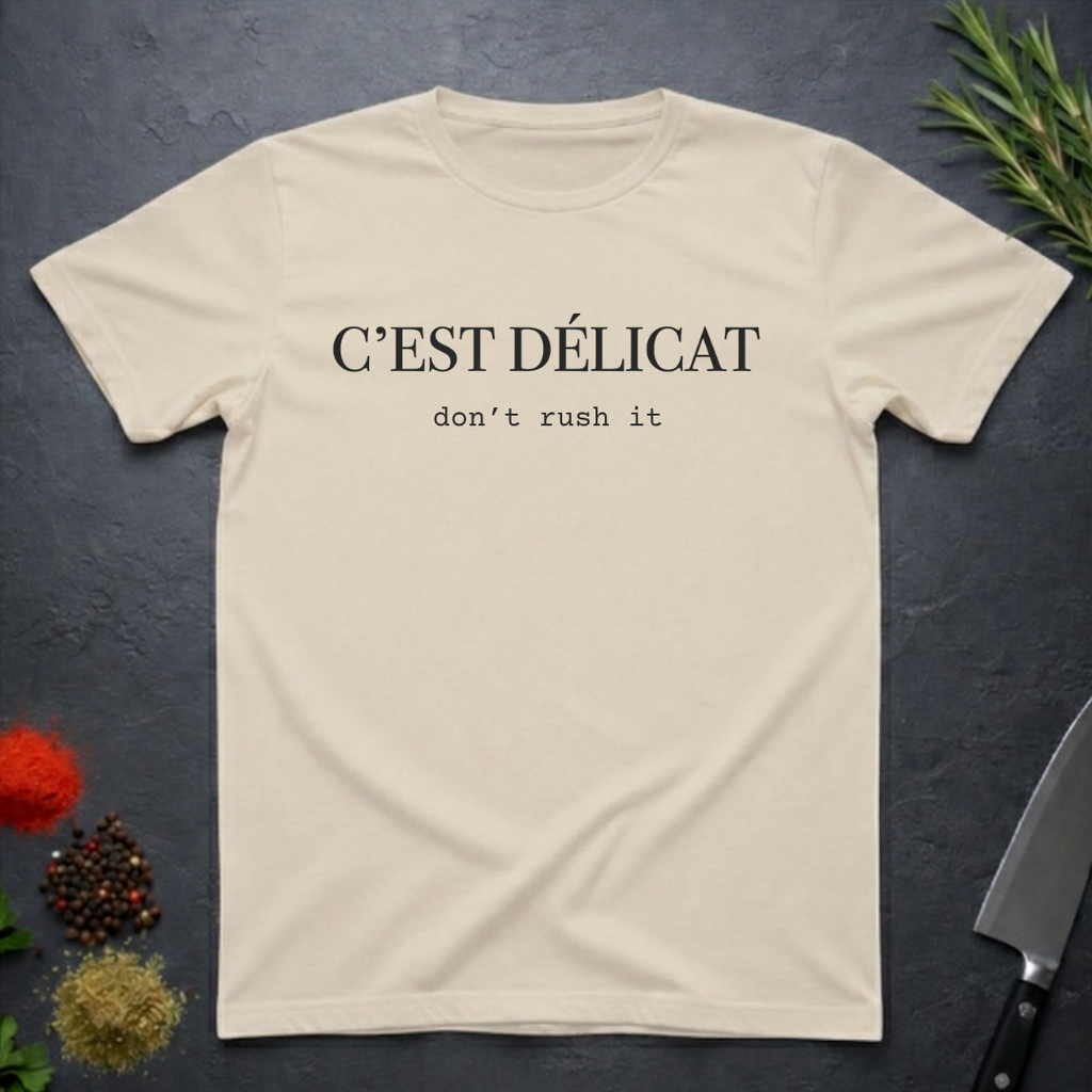 Sand colored t-shirt with text 'C'EST Délicat don't rush it' on a dark surface with decorative elements.
