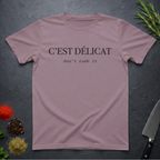 Paragon colored t-shirt with text 'C'EST Délicat don't rush it' on a dark surface with decorative elements.