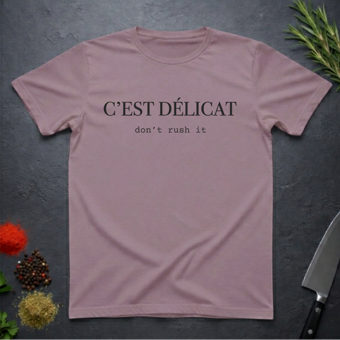Paragon colored t-shirt with text 'C'EST Délicat don't rush it' on a dark surface with decorative elements.