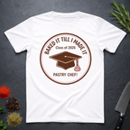 White t-shirt with a Pastry Chef "Baked It Till I Made It - Class of 2026" design on a dark surface with kitchen items.