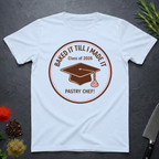 Light Blue t-shirt with a Pastry Chef "Baked It Till I Made It - Class of 2026" design on a dark surface with kitchen items.