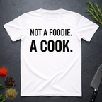 White t-shirt with 'Not a foodie. A cook.' in bold text on a dark surface with kitchen items.