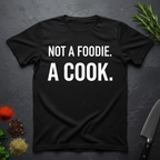 Black t-shirt with 'Not a foodie. A cook.' in bold text on a dark surface with kitchen items.