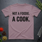 Paragon colored t-shirt with 'Not a foodie. A cook.' text on a dark surface with kitchen items.