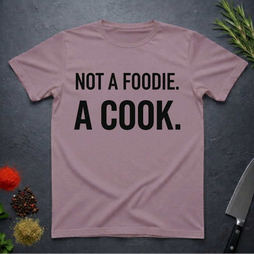 Paragon colored t-shirt with 'Not a foodie. A cook.' text on a dark surface with kitchen items.