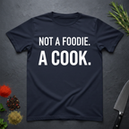 Navy blue t-shirt with 'Not a foodie. A cook.' in bold text on a dark surface with kitchen items.