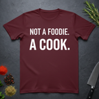 Maroon t-shirt with 'Not a foodie. A cook.' in bold text on a dark surface with kitchen items.