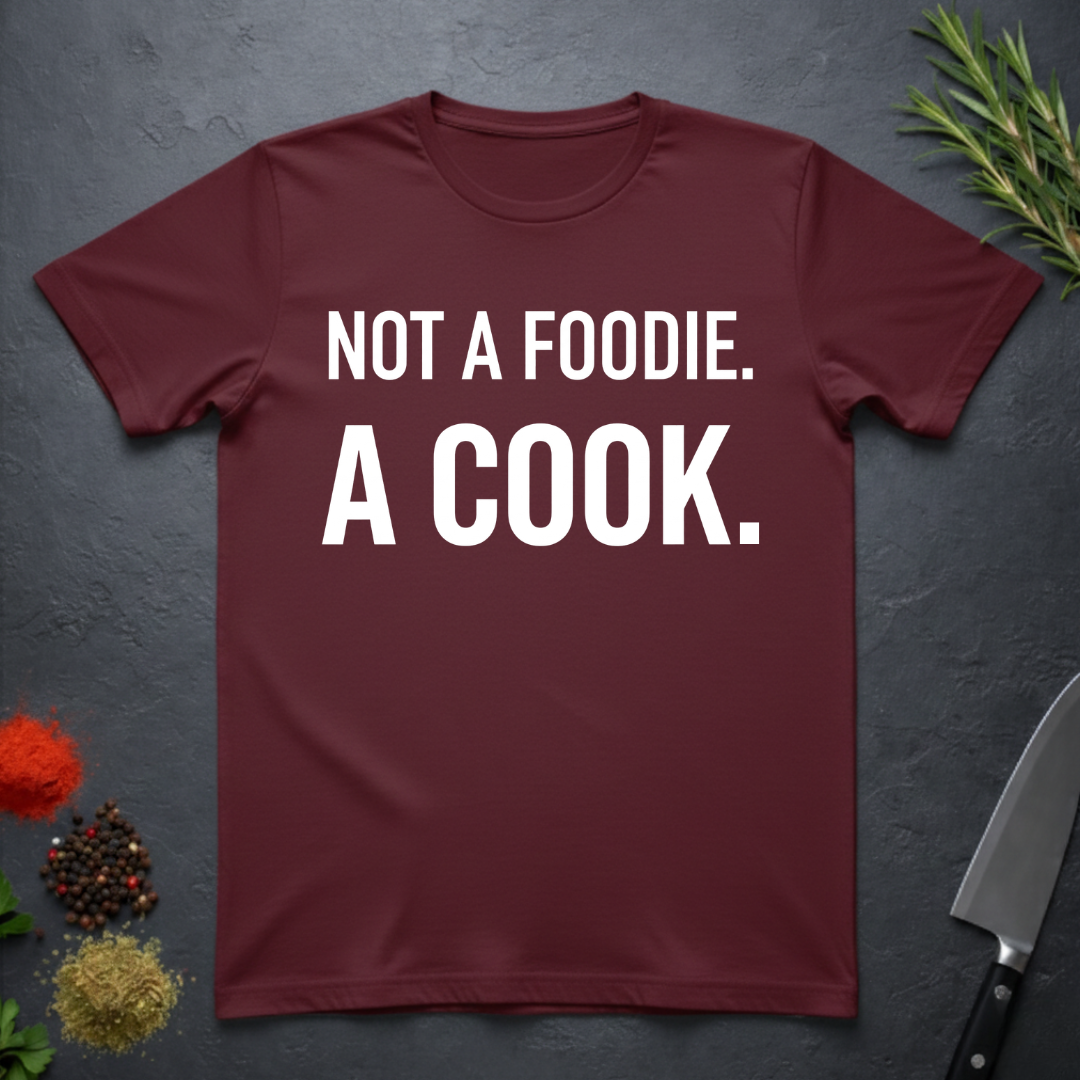Maroon t-shirt with 'Not a foodie. A cook.' in bold text on a dark surface with kitchen items.