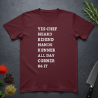 Maroon t-shirt with list of back of house kitchen words in white text on a dark surface with kitchen items.