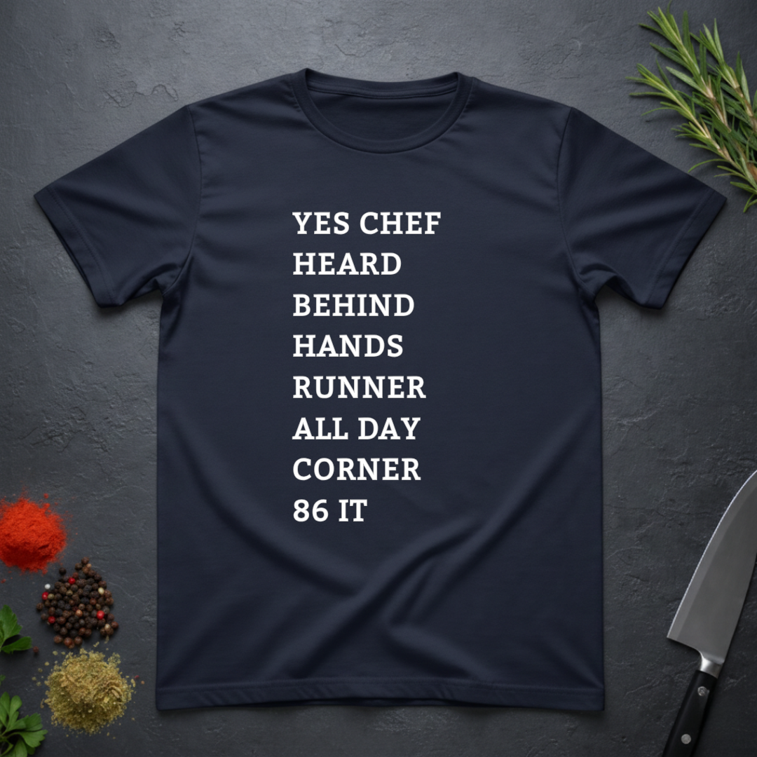 Navy blue t-shirt with list of back of house kitchen words in white text on a dark surface with kitchen items.