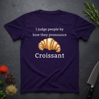 Amethyst purple t-shirt with a croissant graphic and text "I judge people by ho they pronounce Croissant" on a dark surface.