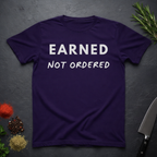 Purple t-shirt with 'Earned Not Ordered' text on a dark surface with decorative elements.
