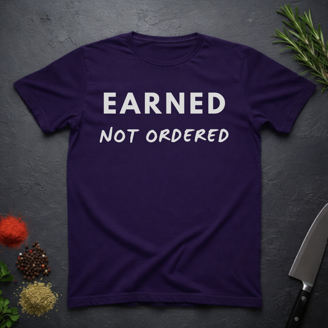Purple t-shirt with 'Earned Not Ordered' text on a dark surface with decorative elements.