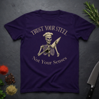 Amethyst purple t-shirt with skeleton design and text "Trust your Steel, Not Your Senses" on a dark surface with decorative elements.
