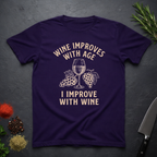 Amethyst purple t-shirt with text "Wine Improves With Age. I Improve With Wine" in an off-white bold font. Image of wine glass and grape bunches separate text. From the Bitter Palate brand.