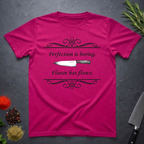 Antique Heliconia pink t-shirt with a knife graphic and text "Perfection is boring. Flavor has flaws." On a dark surface. 