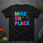 Black t-shirt with colorful fractured 'MISE EN PLACE' text on a dark surface with kitchen items.