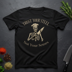 Black t-shirt with skeleton design and text "Trust your Steel, Not Your Senses" on a dark surface with decorative elements.