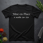 Black t-shirt with 'Mise en Place a motto for life' text on a dark surface with kitchen items.