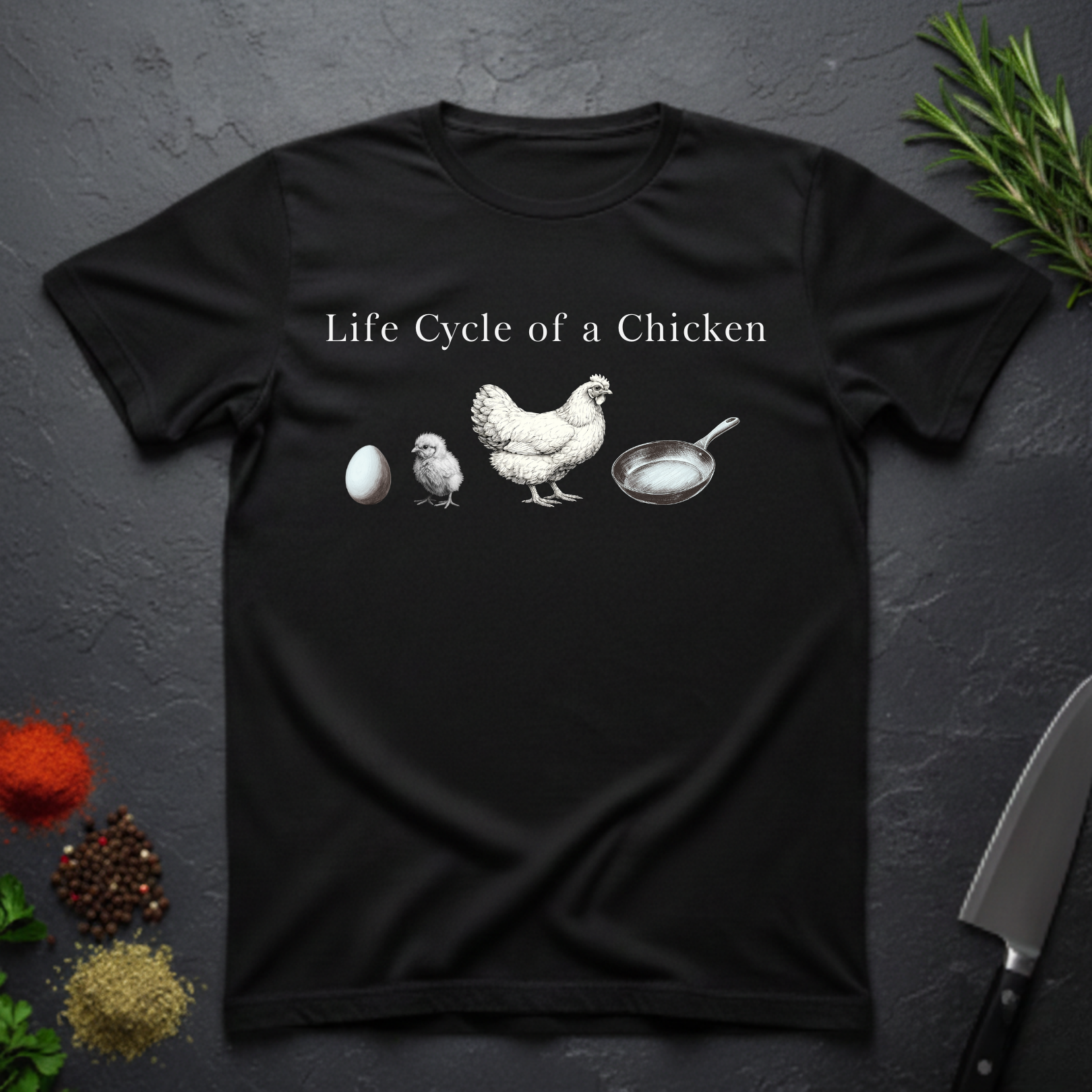 Black t-shirt with text 'Life Cycle of a Chicken' and graphic of an egg, chick, hen, frying pan on a dark surface.