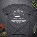 Charcoal grey t-shirt with a knife graphic and text "Perfection is boring. Flavor has flaws." On a dark surface. 